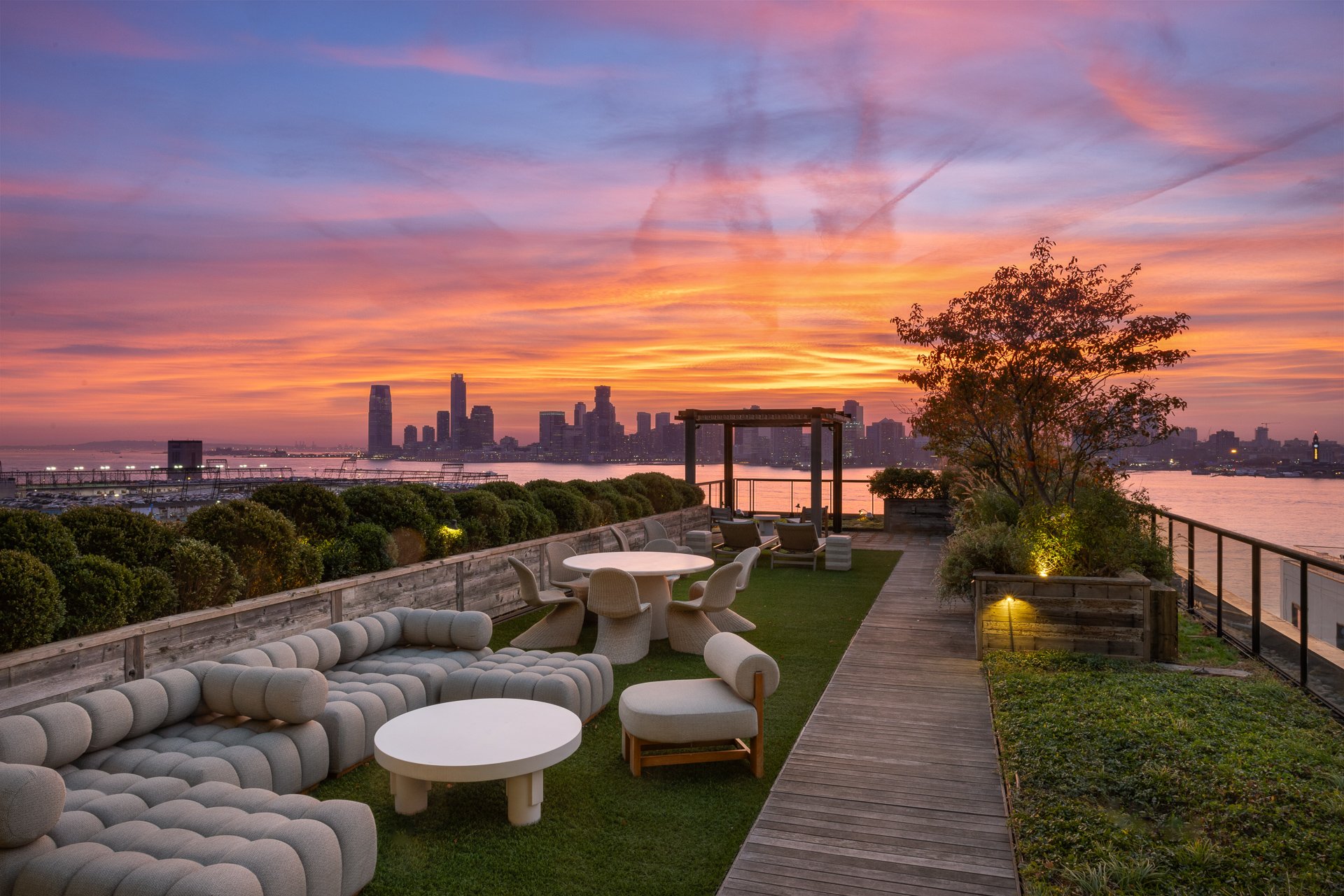 Rooftop lounge with seating area including a large sofa, chairs, and tables, overlooking a city skyline at sunset with colorful sky and water in the background.
