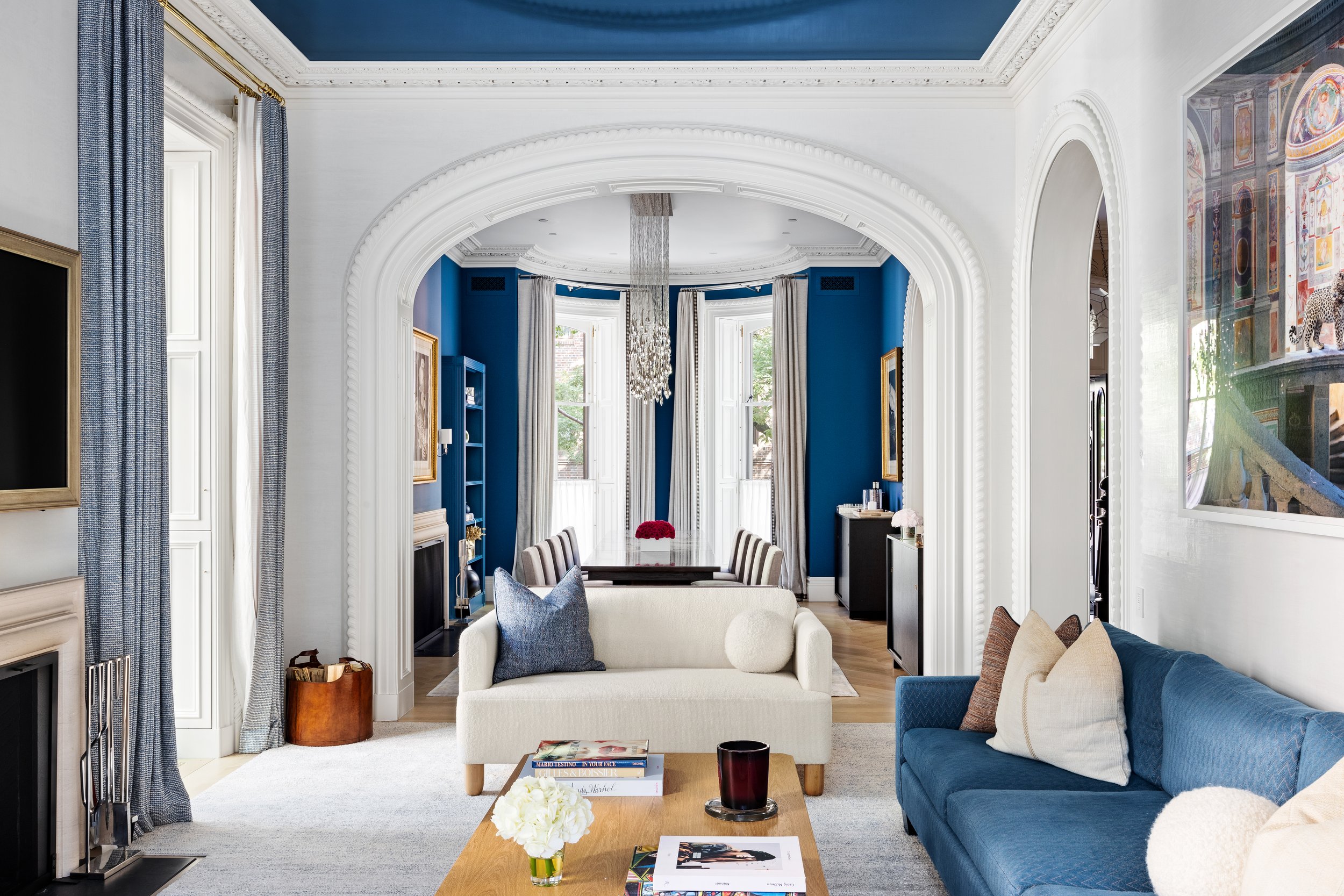 Living room with blue and white color scheme, featuring a white sofa, a blue armchair, a wooden coffee table with books and a candle, and a fireplace. In the background, a dining area with a dark wood table, beige chairs, and a chandelier, with blue walls and white curtains in windows.