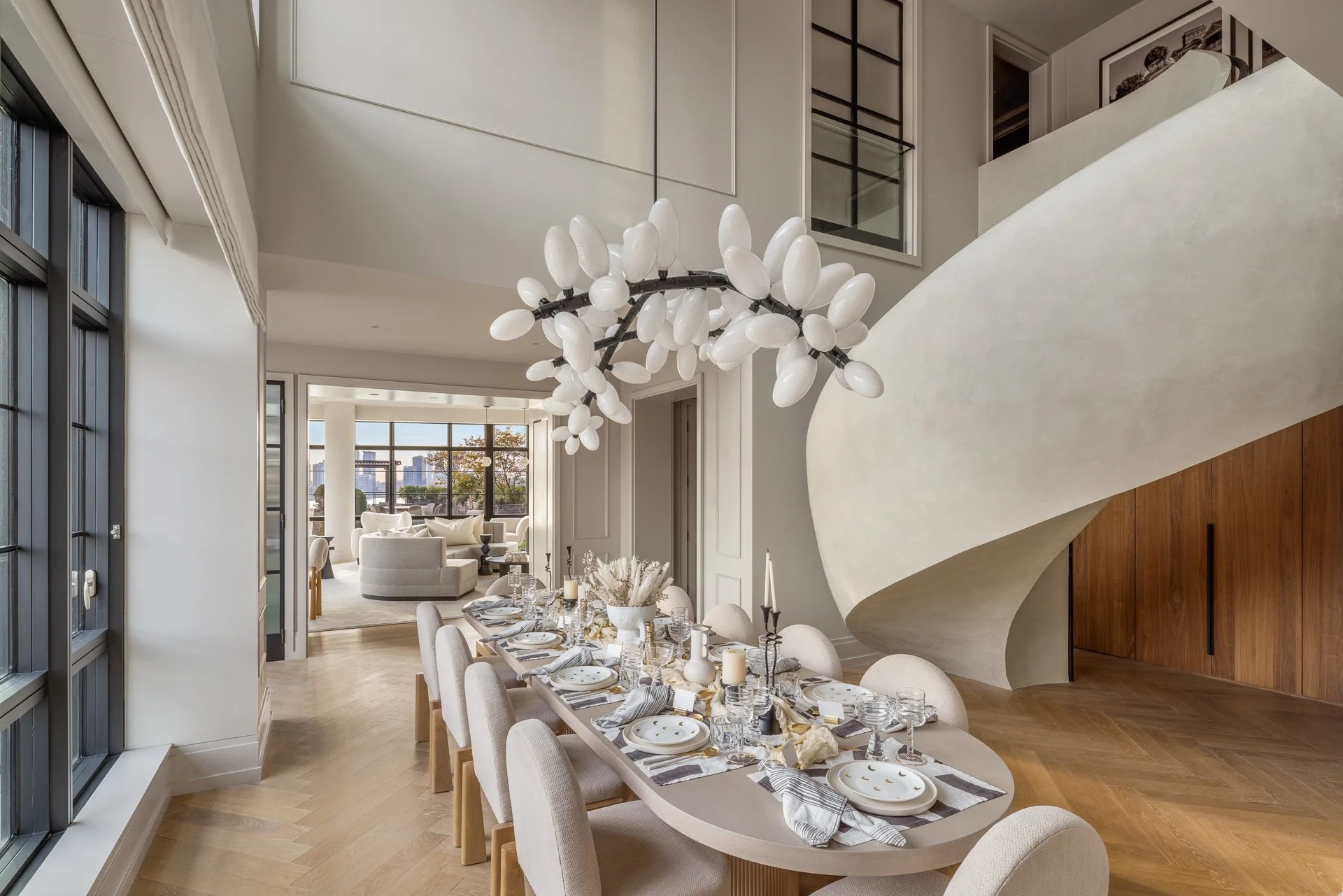 Elegant modern dining room with a long table set for a meal, a white sculptural chandelier, and an open living area with large windows in the background