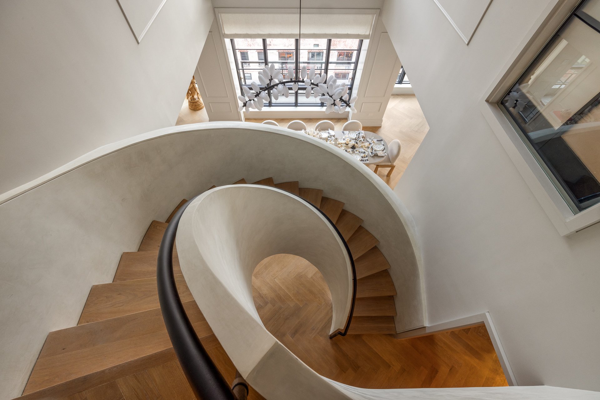 View of a spiral staircase from above in a modern home, with a dining area visible at a lower level through large windows.