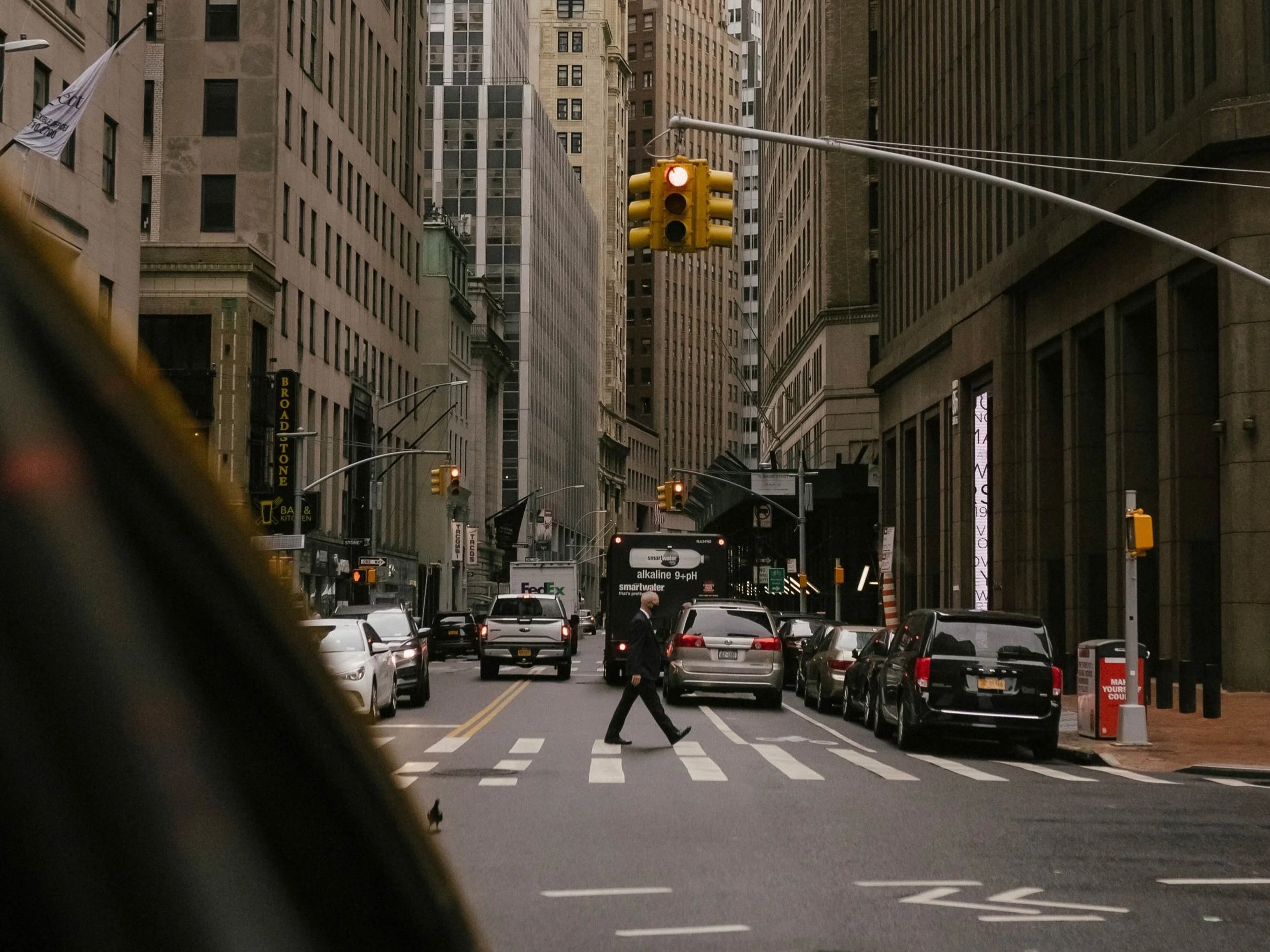 City street scene with tall buildings, cars parked along the sides, a person walking across the crosswalk, and a yellow traffic light.