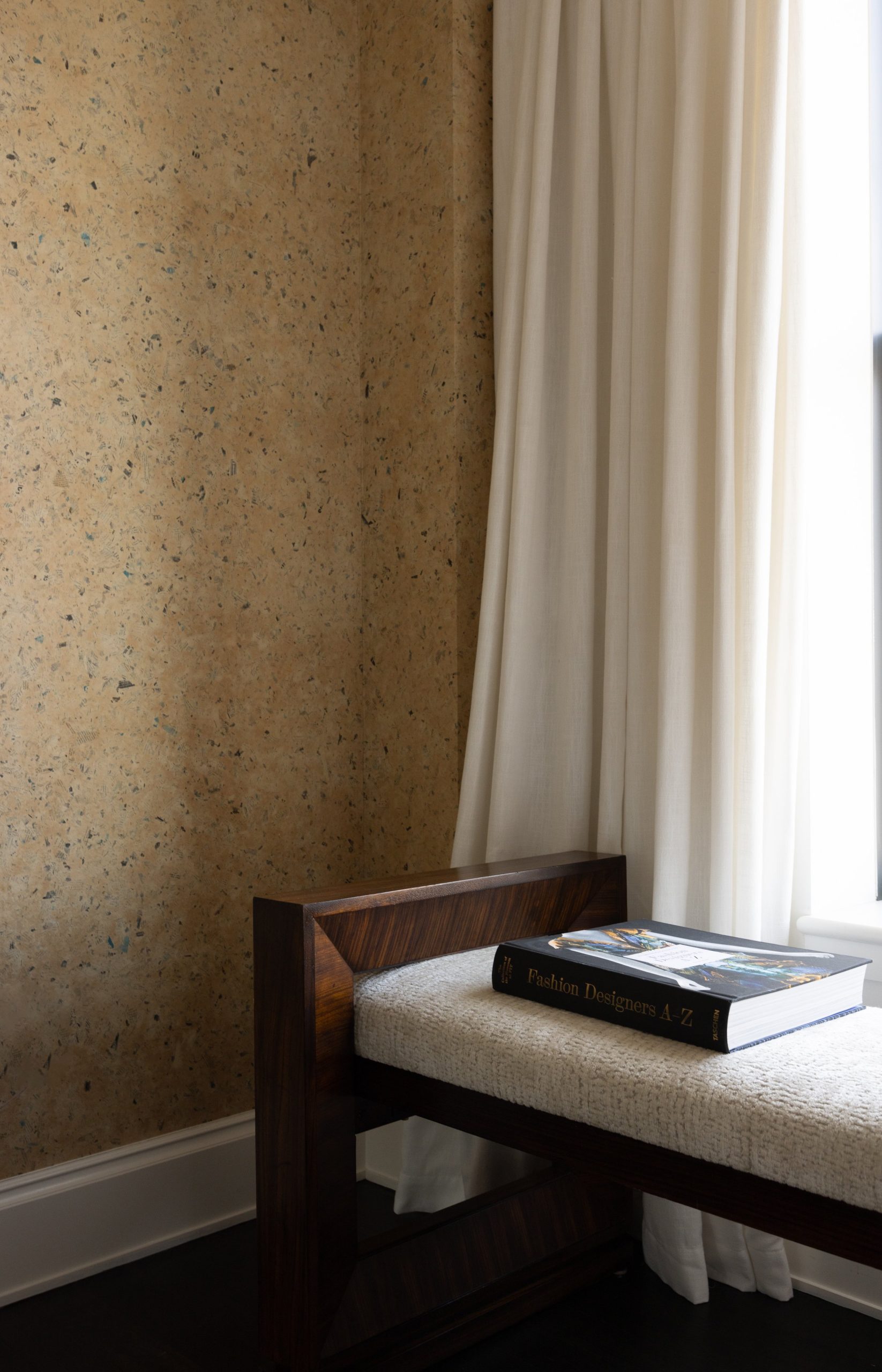 A corner of a room with a textured beige wall, white curtains, and a wooden bench with a book titled 'Fashion Designers A-Z' on top.
