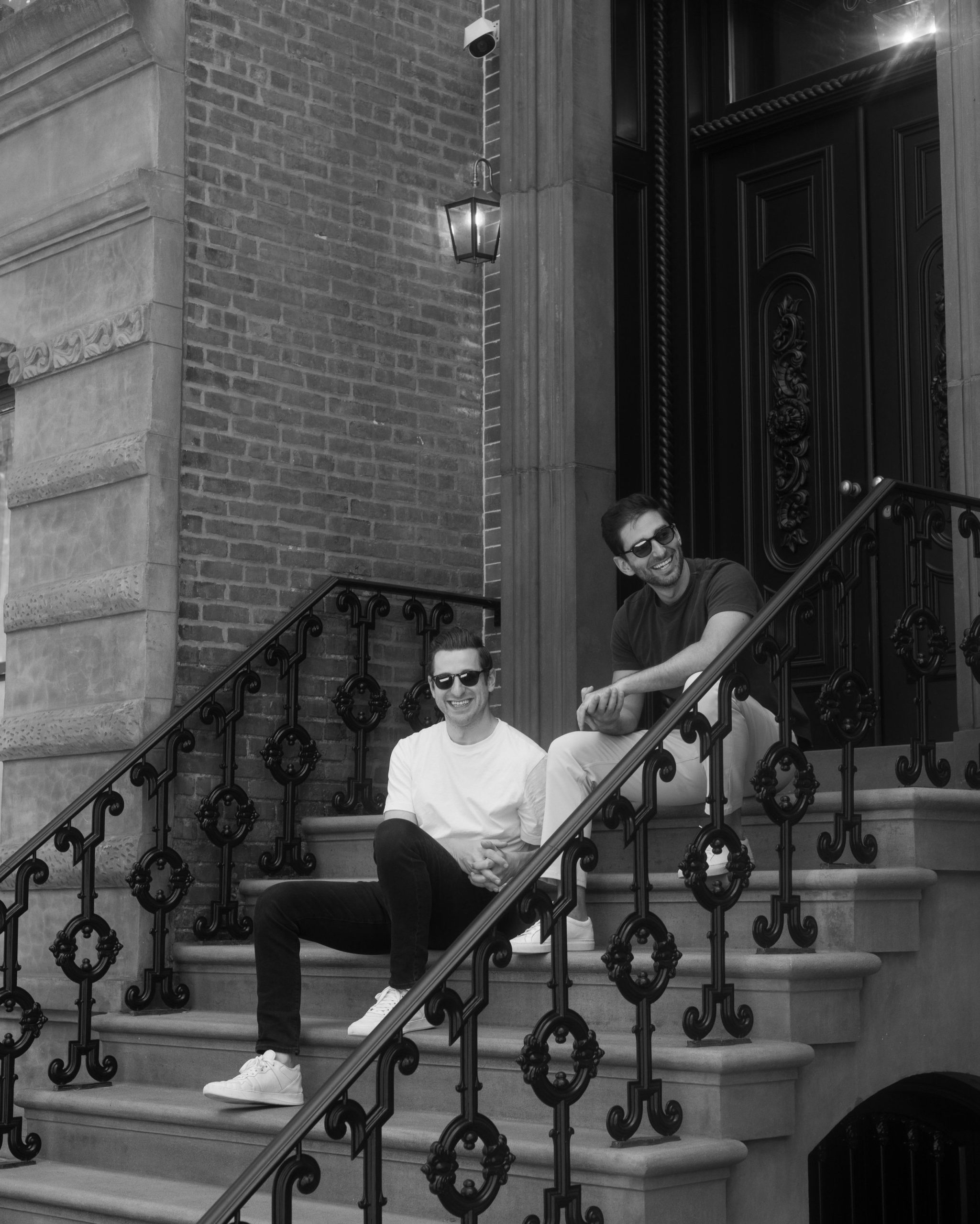 Two men wearing sunglasses sitting on the steps of a building, smiling and looking relaxed, with ornate black railings and a brick wall behind them.