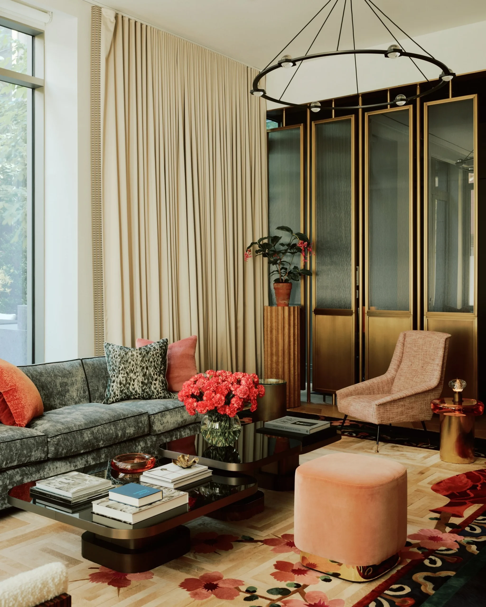 A Manhattan living room: full height curtains beside a wall of glazed brass cabinets, a plant on a fluted pedestal and an upholstered armchair on a herringbone floor.