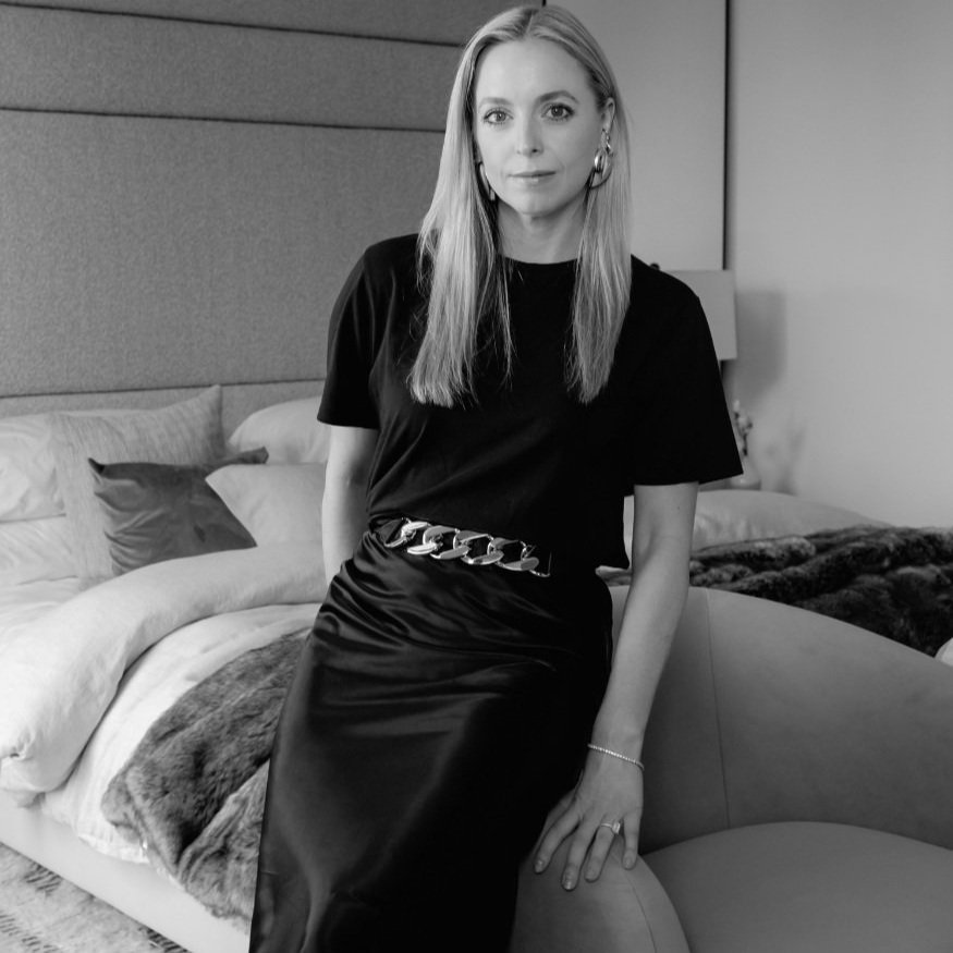 A woman with long hair wearing a black dress and accessories, sitting on a upholstered armchair in a bedroom with bed and pillows in the background