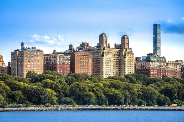 View of a city skyline with tall buildings and lush green trees in front, with a body of water in the foreground and a clear blue sky.