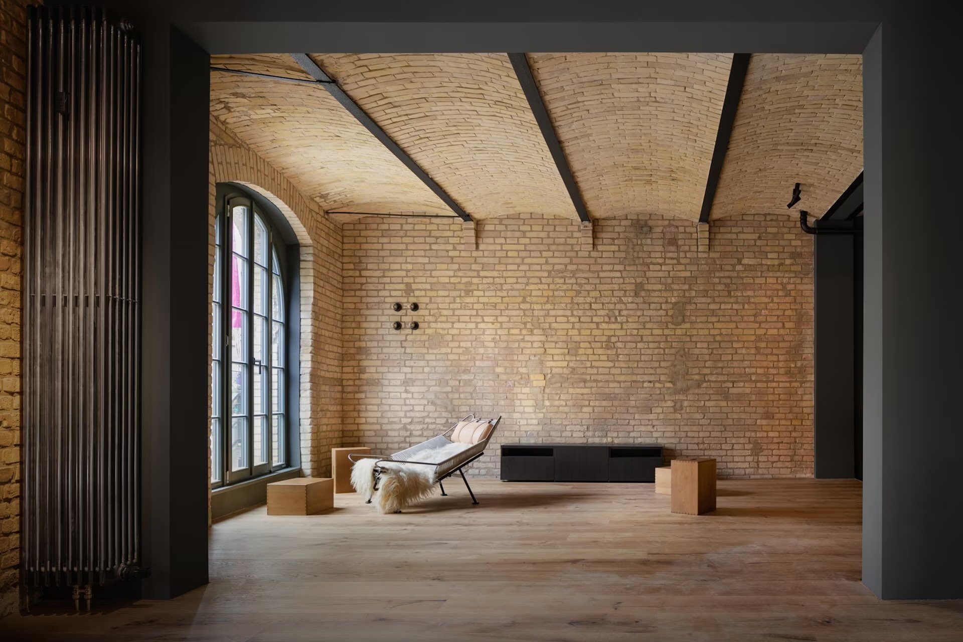 An interior space with exposed brick walls, wooden flooring, a large arched window, and a black modern lounge chair with a sheepskin throw, surrounded by minimalist wooden furnishings.